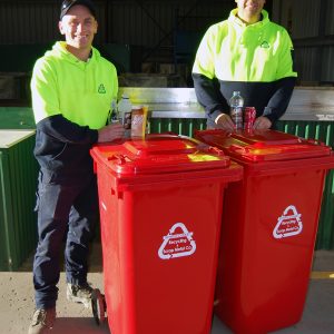 Andrew and Mark in hi-vis yellow jumpers with bottles and cans and the red wheelie bins with South Coast Recycling logo in white for the new bottle and can collection service.